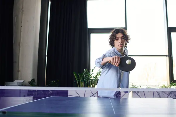 Attractive young man plays ping pong in bright office environment with large windows and greenery. — Stock Photo