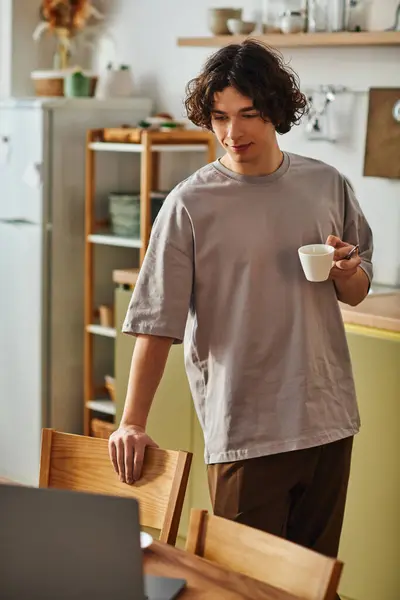 A young attractive man stands in a modern office, savoring coffee as he glances at his laptop. — Stock Photo