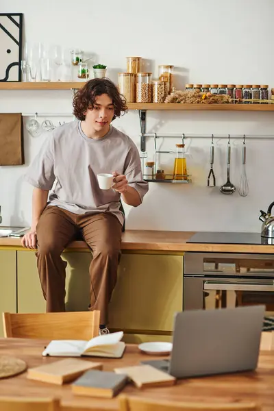 A handsome young man sips coffee on a countertop, gazing at his workspace and notes. — Stock Photo