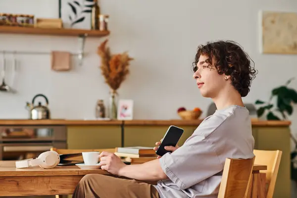 Young man sits thoughtfully at a wooden table with a phone and coffee in a bright office. — Stock Photo