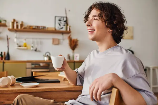 A young man with curly hair smiles while sipping coffee in a chic office, exuding positivity. — Stock Photo