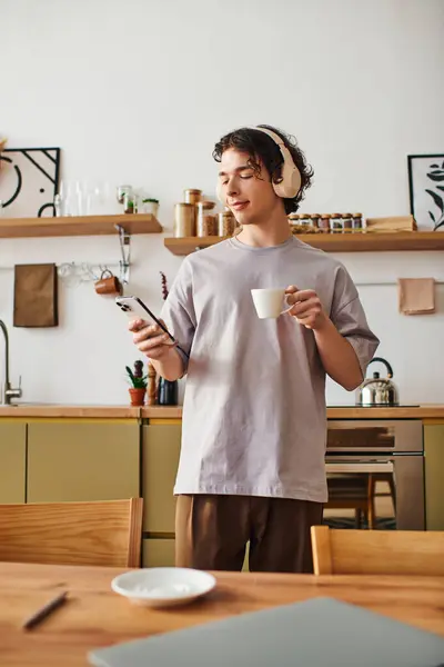 Handsome young man wearing headphones sips coffee and checks his phone in a stylish office. — Stock Photo