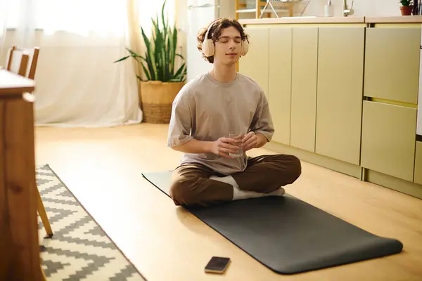 Young man with headphones relaxes on a mat in a sunny office, sipping a drink and meditating. — Stock Photo