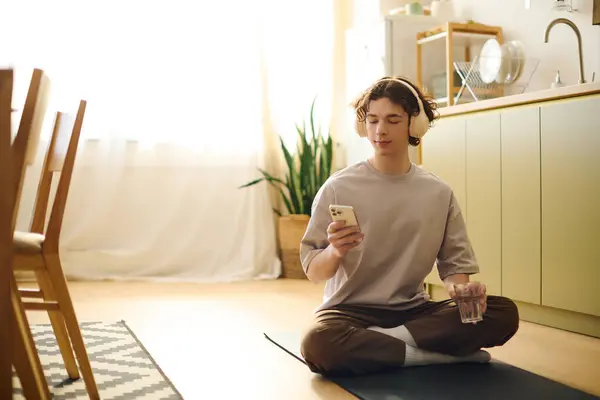 A young man sits in a comfortable position, focused on his phone while sipping water. — Stock Photo