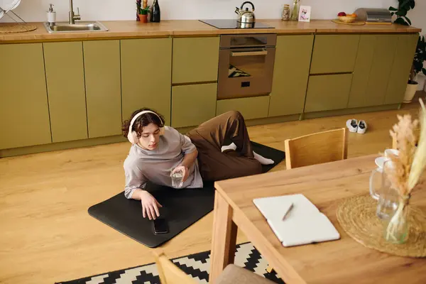A handsome young man lounges on the floor with headphones, sipping water and using his phone. — Stock Photo