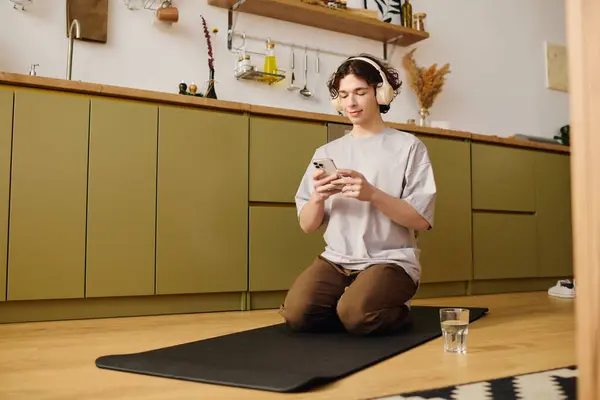 Un giovane uomo si gode un momento di relax mentre usa il suo telefono, seduto su un tappeto in una cucina accogliente. — Stock Photo