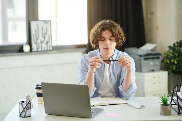 A young man sits in an office, pen in hand, deep in thought with a laptop and notebook. — Stock Photo