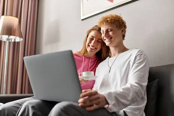A young couple bonds over a laptop in their hotel room, sharing smiles and warm drinks together. — Stock Photo
