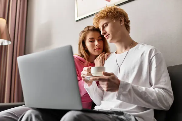 A couple cuddles on a hotel couch, sharing smiles and a warm drink while on a laptop. — Stock Photo