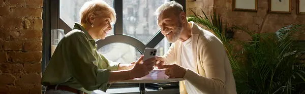 A middle-aged couple smiles and shares a moment while looking at their phone at a cafe. — Stock Photo