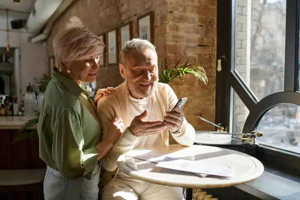 A middle aged couple share laughter and joy over a smartphone at a charming cafe. — Stock Photo