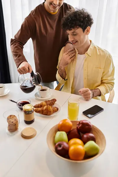 Pareja gay disfrutar de un delicioso desayuno como uno vierte café para el otro, rodeado de frutas frescas. - foto de stock