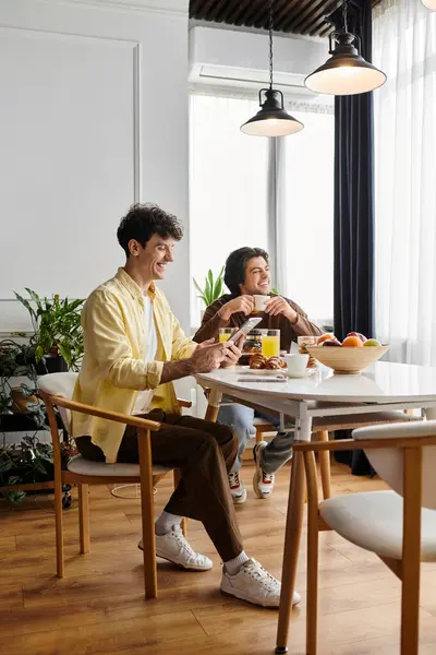 Due uomini condividono una mattinata gioiosa nel loro elegante appartamento, gustando la colazione e sorridendo.. — Foto stock