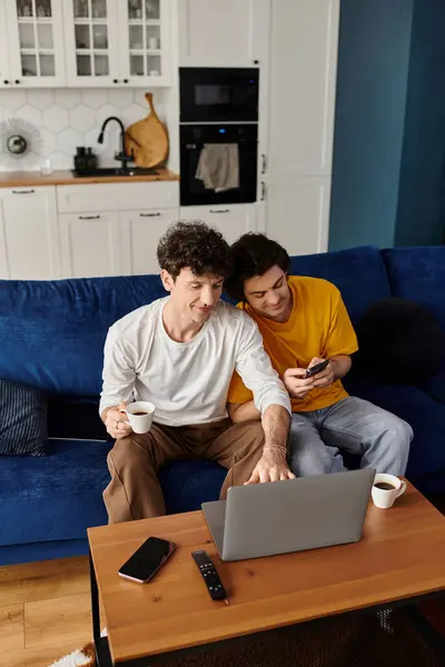 Due uomini condividono un momento gioioso nel loro appartamento moderno mentre usano un computer portatile e si godono il caffè. — Foto stock