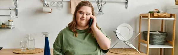 A young pretty woman with Down syndrome sits in a cozy kitchen, chatting comfortably on the phone. — Stock Photo