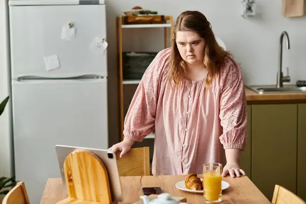 In a cozy kitchen, a young woman with Down syndrome enjoys breakfast while using her laptop. — Stock Photo