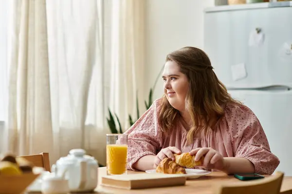 A young woman with Down syndrome enjoys a croissant and juice at a cozy table. — Stock Photo