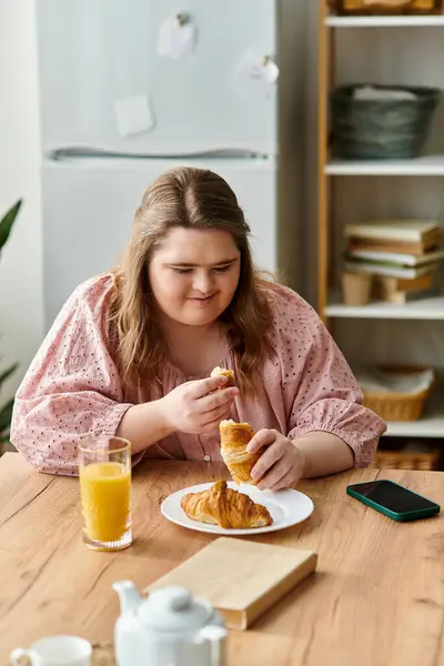 Young woman with Down syndrome sits in a comfy home environment, savoring a croissant and juice. — Stock Photo