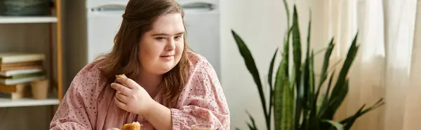 A pretty young woman with Down syndrome sits comfortably at home, savoring a snack with joy. — Stock Photo