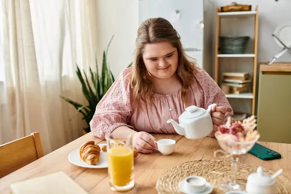 Una joven con síndrome de Down vierte con alegría el té en casa mientras disfruta de un croissant. - foto de stock