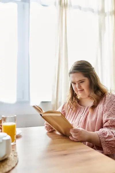 In a sunny home setting, a young woman reads a book, savoring a refreshing glass of juice. — Stock Photo