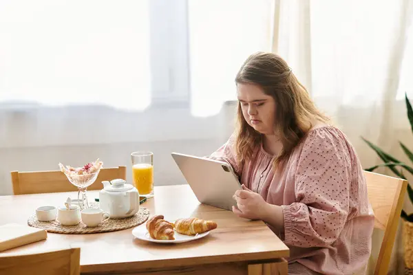A young woman with Down syndrome sits at a wooden table, enjoying breakfast on her tablet. — Stock Photo
