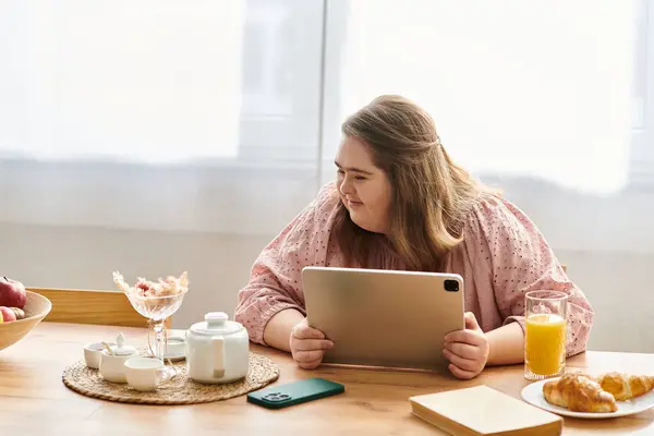 A young woman with Down syndrome smiles at a breakfast table with a tablet. — Stock Photo
