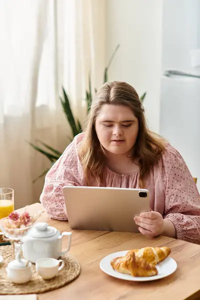 A young woman with Down syndrome relaxes at home while using a tablet, enjoying a light breakfast. — Stock Photo