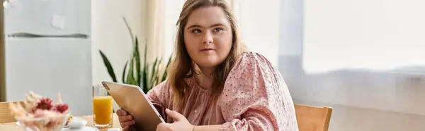 Giovane donna con sindrome di Down siede a un tavolo, godendo di succo e utilizzando una tavoletta. — Foto stock