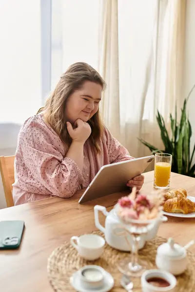 Une jeune femme trisomique prend son petit déjeuner à la maison en utilisant une tablette. — Photo de stock