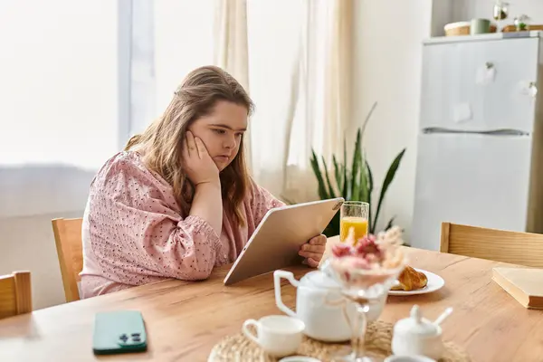 Una joven con síndrome de Down se sienta en una mesa de comedor, comprometida con su tableta. - foto de stock