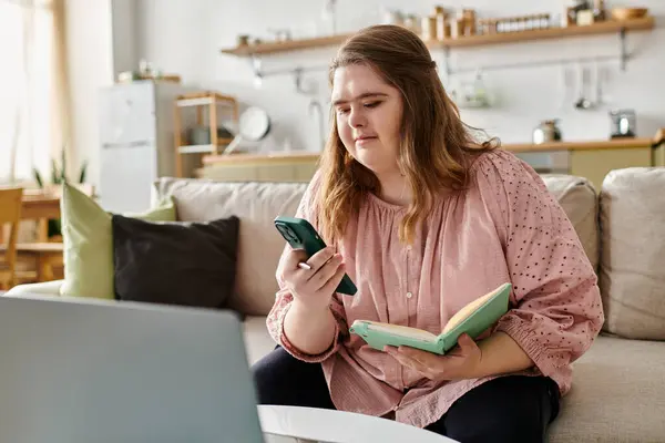 A young woman with Down syndrome enjoys a cozy moment at home with her phone and a book. — Stock Photo