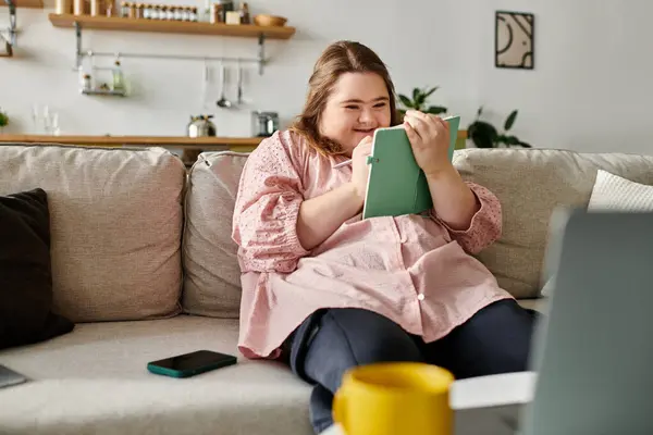 A young woman with Down syndrome sits on a sofa, joyfully writing in her notebook at home. — Stock Photo