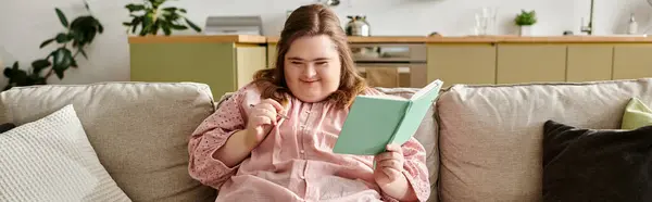 Uma jovem mulher bonita com síndrome de Down senta-se em um sofá, lendo um livro e sorrindo alegremente. — Fotografia de Stock