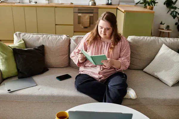In a cozy home, a young woman with Down syndrome sits on a sofa, focused on her book. — Stock Photo