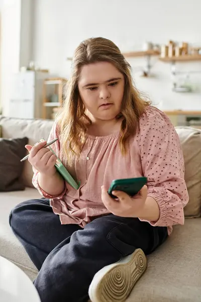 Jolie jeune femme trisomique s'engage avec son téléphone dans un espace de vie détendu et confortable. — Photo de stock