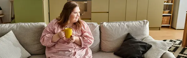 Young woman with Down syndrome relaxes on a comfy couch, sipping from a cheerful yellow mug. — Stock Photo