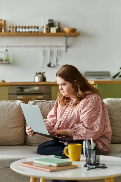 A pretty young woman with Down syndrome sits comfortably on a sofa, focused on her laptop at home. — Stock Photo