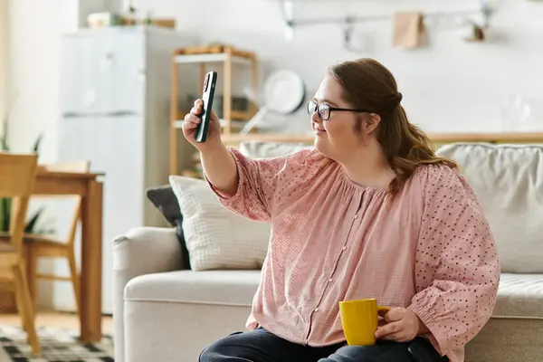Una joven con síndrome de Down sonríe en casa, sosteniendo su teléfono y café. - foto de stock