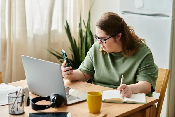 Young woman with Down syndrome sits at her desk, using her phone and notebook. — Stock Photo