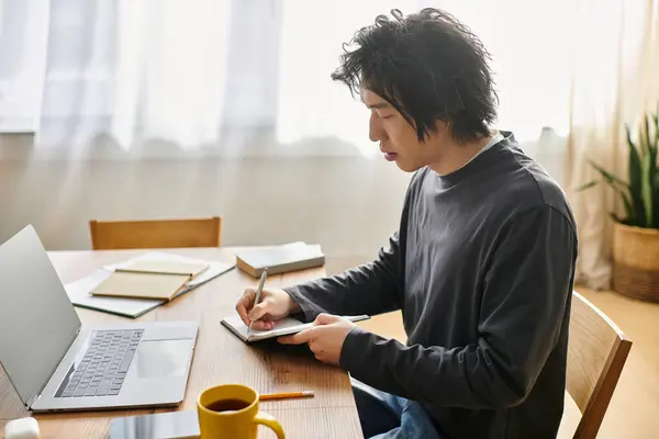 A young man is diligently studying online from his home, taking notes while focused on his laptop. — Stock Photo