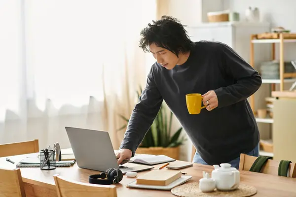 A handsome young Asian man engages in online studies while holding a warm beverage at his desk. — Stock Photo