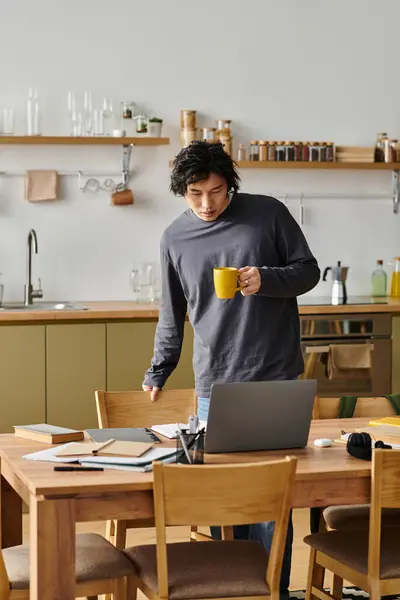 Handsome young man immersed in his online studies while enjoying a warm drink at home. — Stock Photo