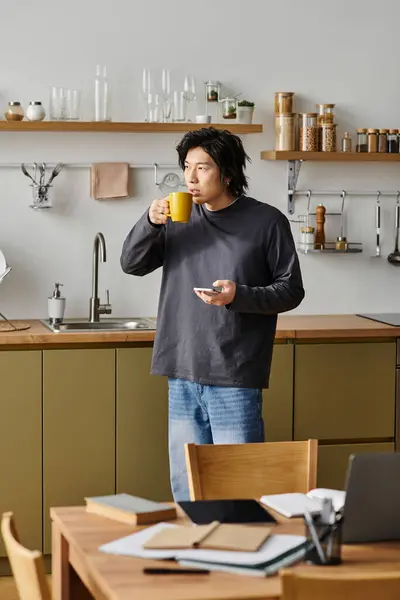 A handsome young Asian man stands in his kitchen, sipping coffee and focusing on his laptop. — Stock Photo