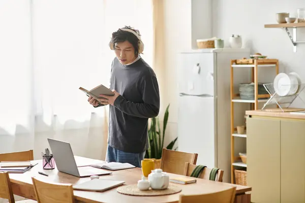 Un joven asiático estudia en línea en casa, centrado en su ordenador portátil y materiales de aprendizaje. - foto de stock