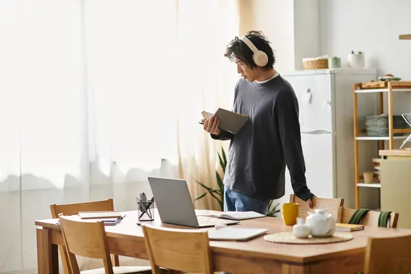 A handsome young man studies online at home with a book in hand and laptop nearby, deeply engaged. — Stock Photo
