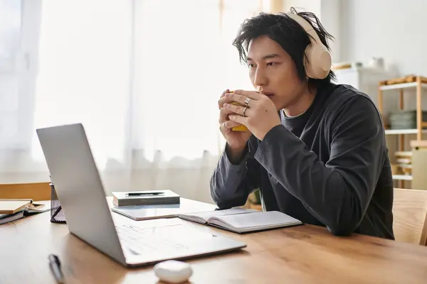 A handsome young asian man is focused on his studies, using a laptop while enjoying a warm drink. — Stock Photo
