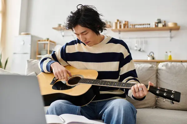 A young Asian man sits on a couch, focused on learning acoustic guitar from a book. — Stock Photo