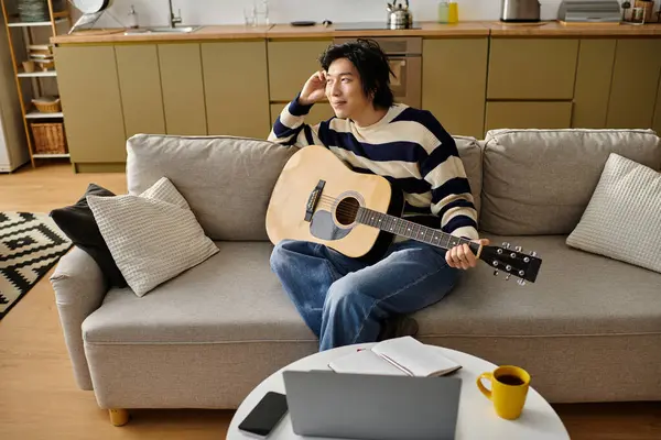 Un giovane uomo si siede rilassato su un divano, praticando la sua chitarra acustica, godendo il momento. — Foto stock