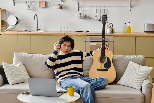 Handsome young Asian man sits comfortably on a couch while practicing guitar, focused and inspired. — Stock Photo
